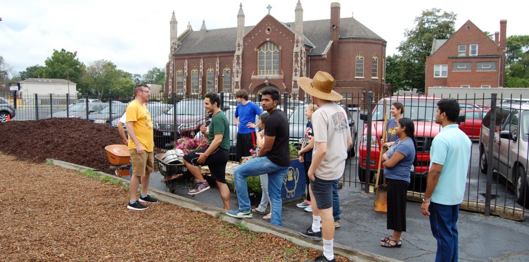 Group of students standing with shovels and rakes outside, in front of a fence near a parking lot, near a large pile of mulch, listening to an individual in a yellow shirt speaking