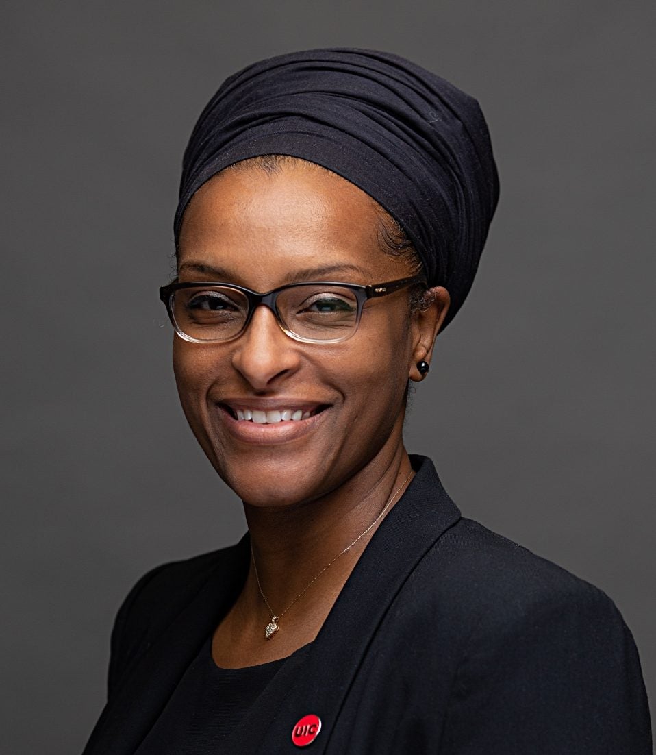 Dr. El-Amin facing the camera smiling in black headwrap and black blazer with red UIC pin